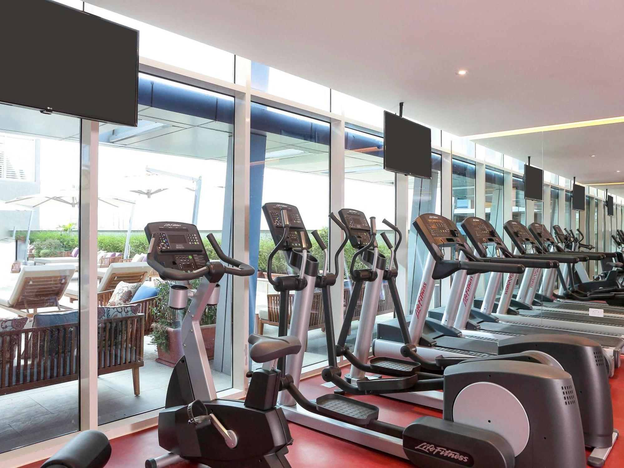 Novotel Jazan Exterior photo The photo shows a modern gym interior with several exercise machines, specifically elliptical trainers, arranged neatly in rows. Large windows allow natural light to fill the space, providing a view outside to a patio area with lounge chairs. There a