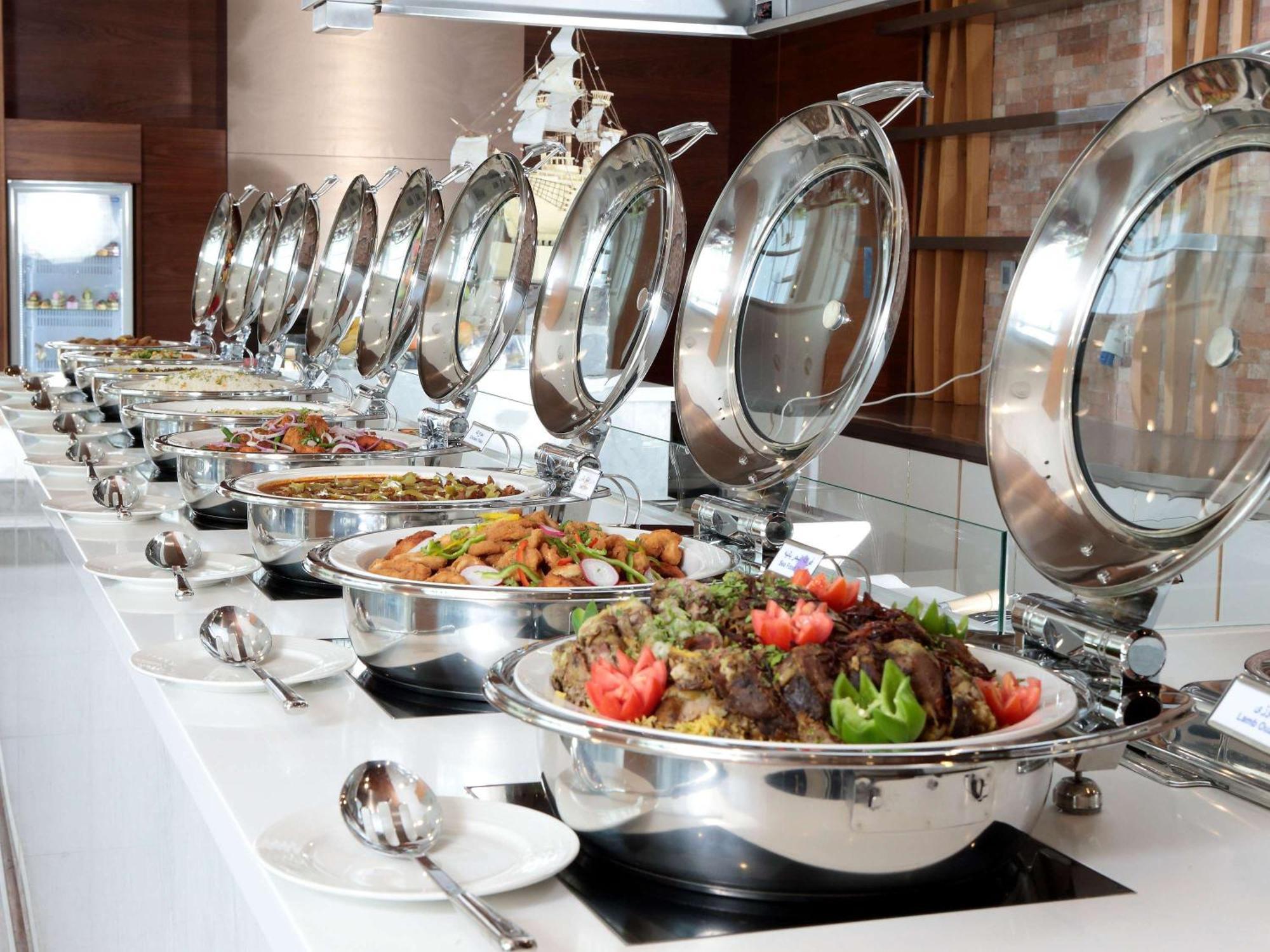 Novotel Jazan Exterior photo The photo shows a buffet setup with a long row of stainless steel chafing dishes. Each dish has a lid, and they are arranged neatly on a white countertop. The food inside appears to be a variety of dishes, including a colorful mix of vegetables and p